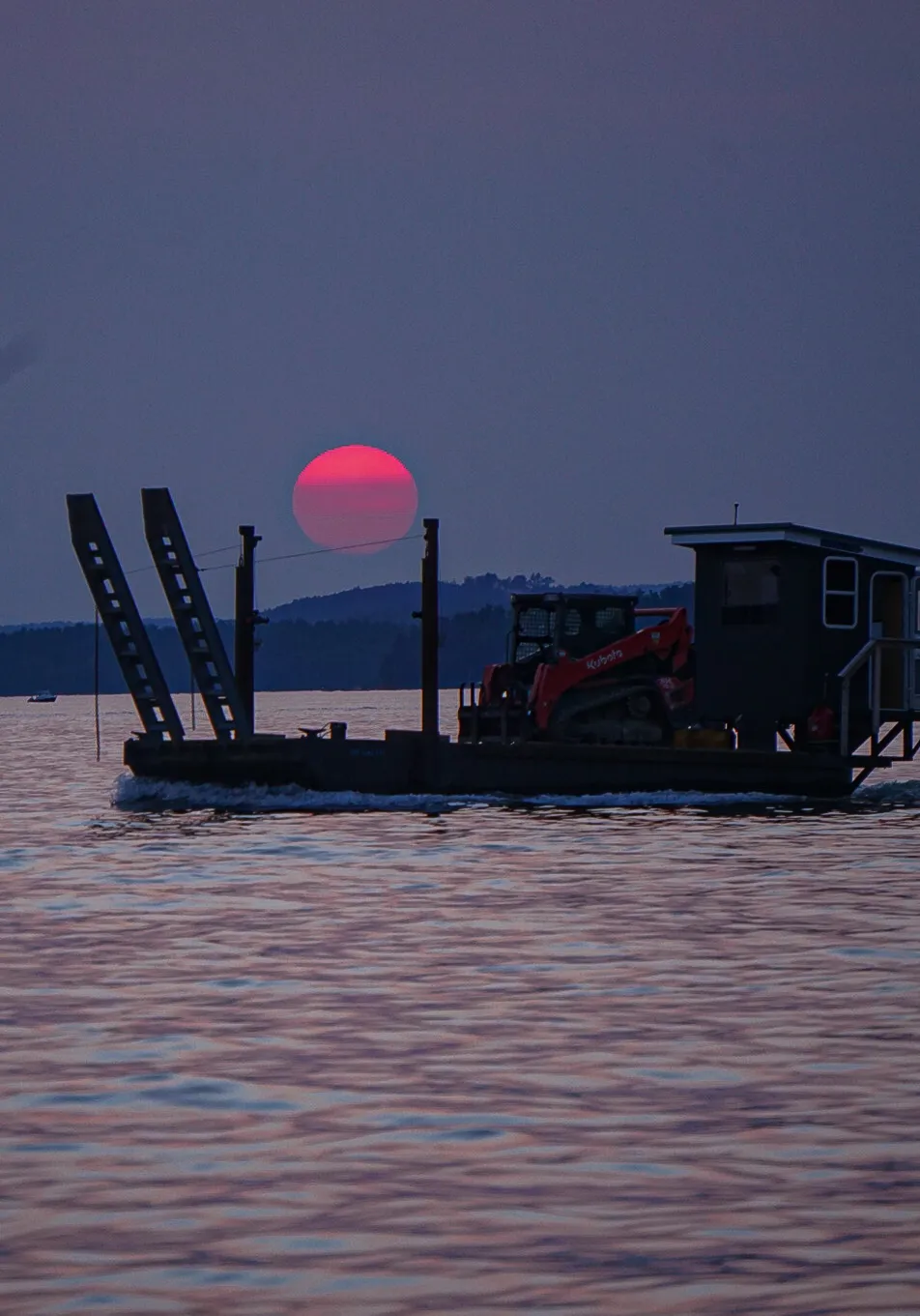 A barge crossing a lake at dusk with a deep red sun setting behind hills