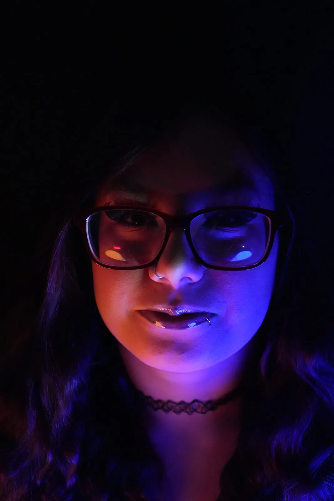 A moody close portrait of a woman in glasses, her face split between warm ember and cool blue light against black